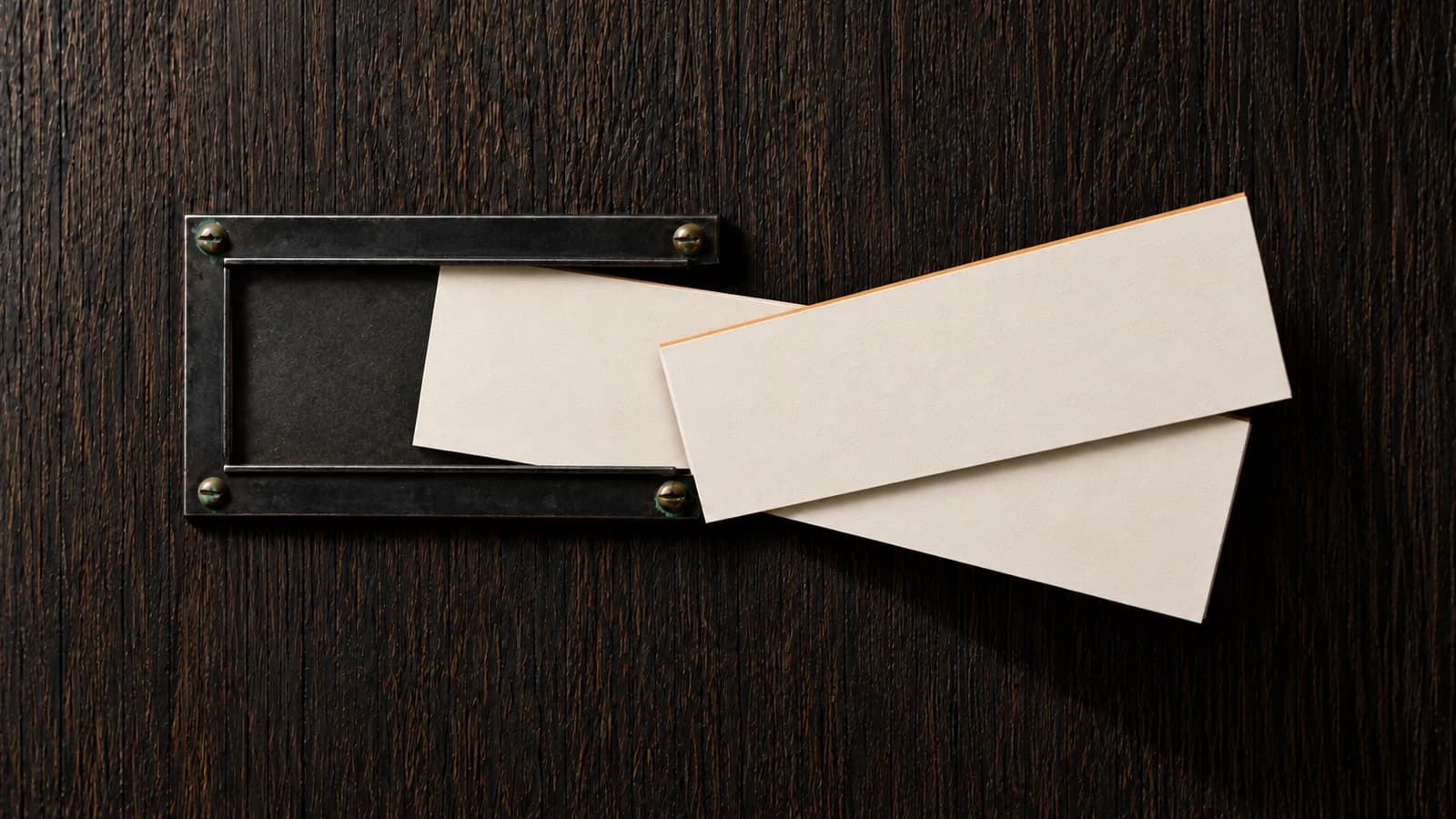 A blank ivory card tilts out of a blackened-steel card frame screwed to a dark oak wall, as a second blank card with burnt-amber edges crosses over it on its way in.