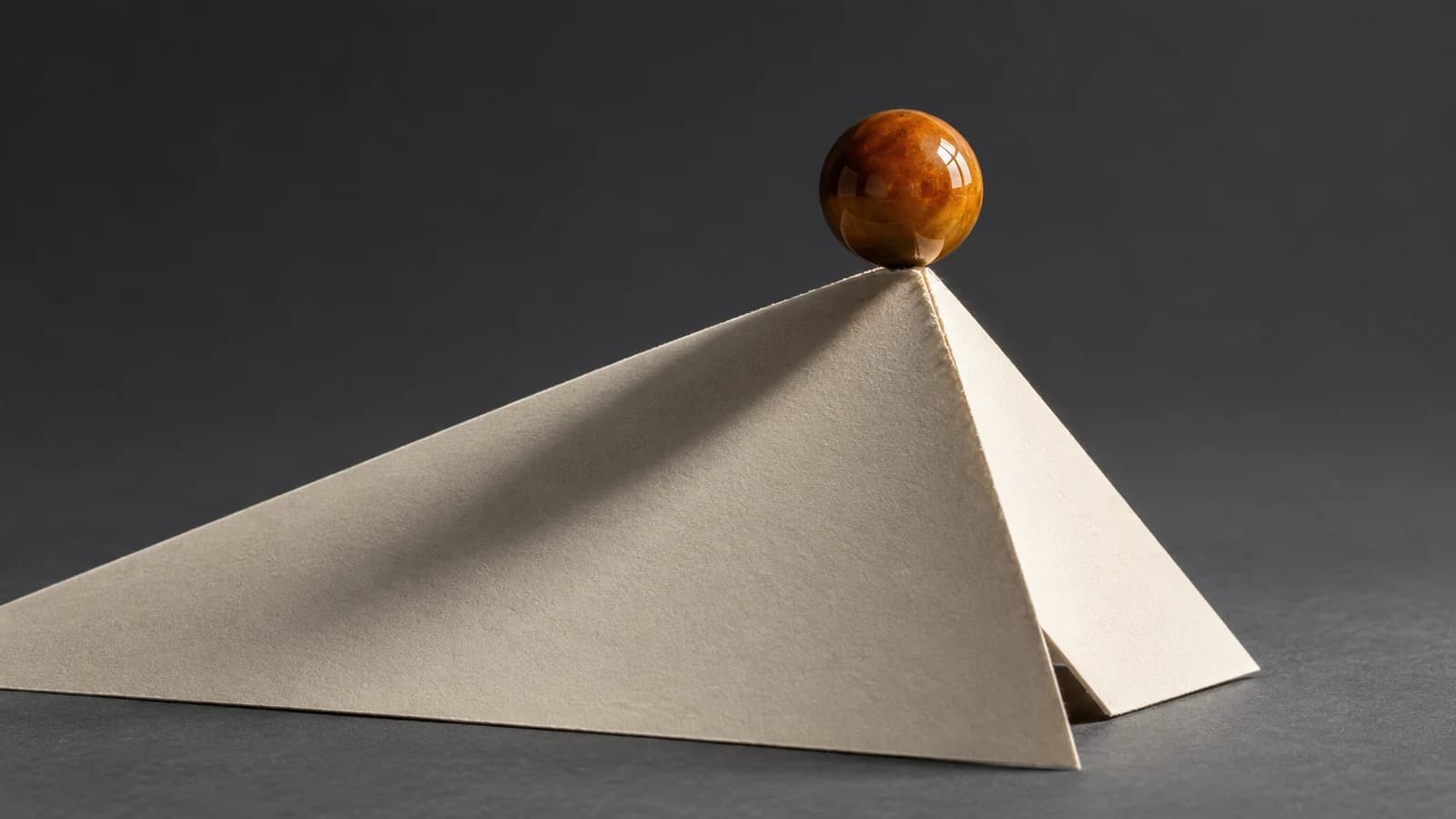 An amber glazed ball balances on the sharp apex of a folded ivory card peak, with a long shallow slope to the left and a short steep face to the right, on a dark smoky-grey ground.